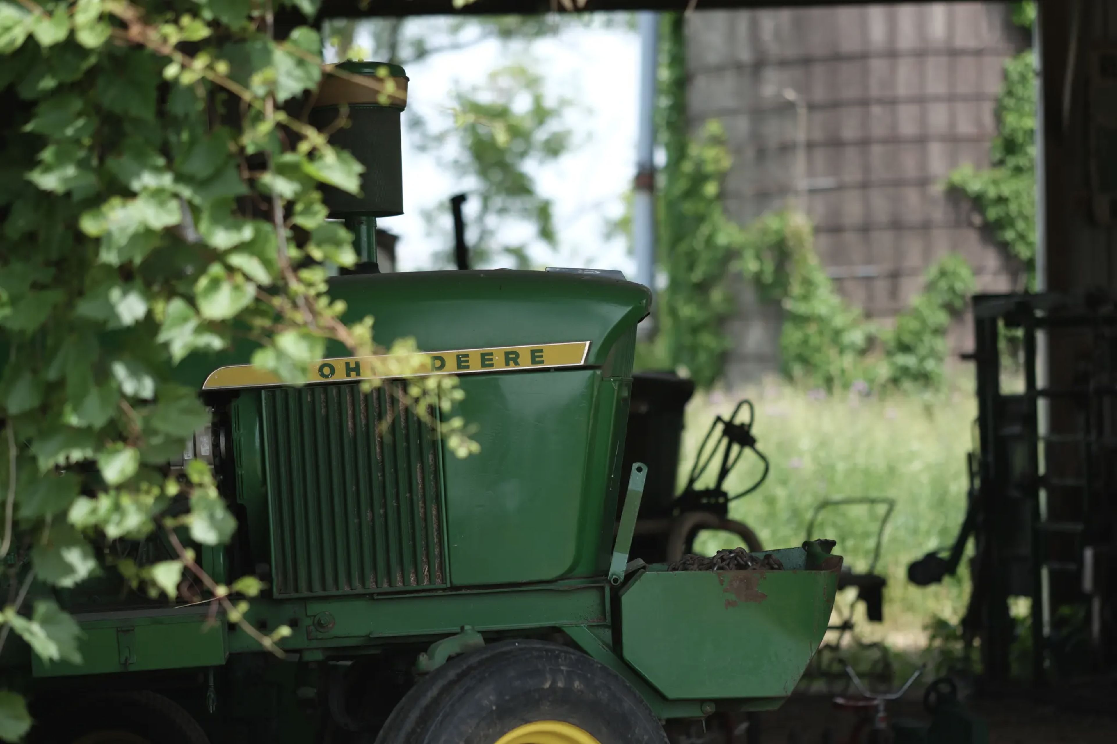 A green tractor partially obscured by foliage, parked in a shaded area with a silo and greenery in the background