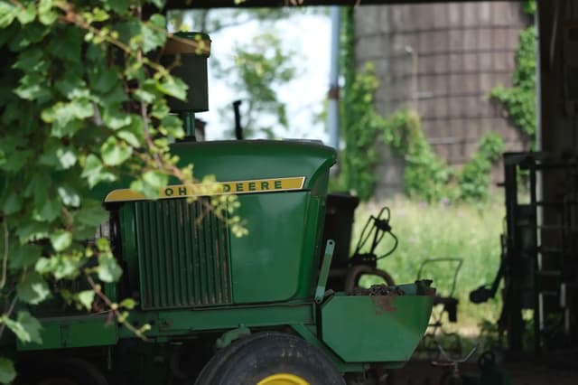 A green tractor partially obscured by foliage, parked in a shaded area with a silo and greenery in the background